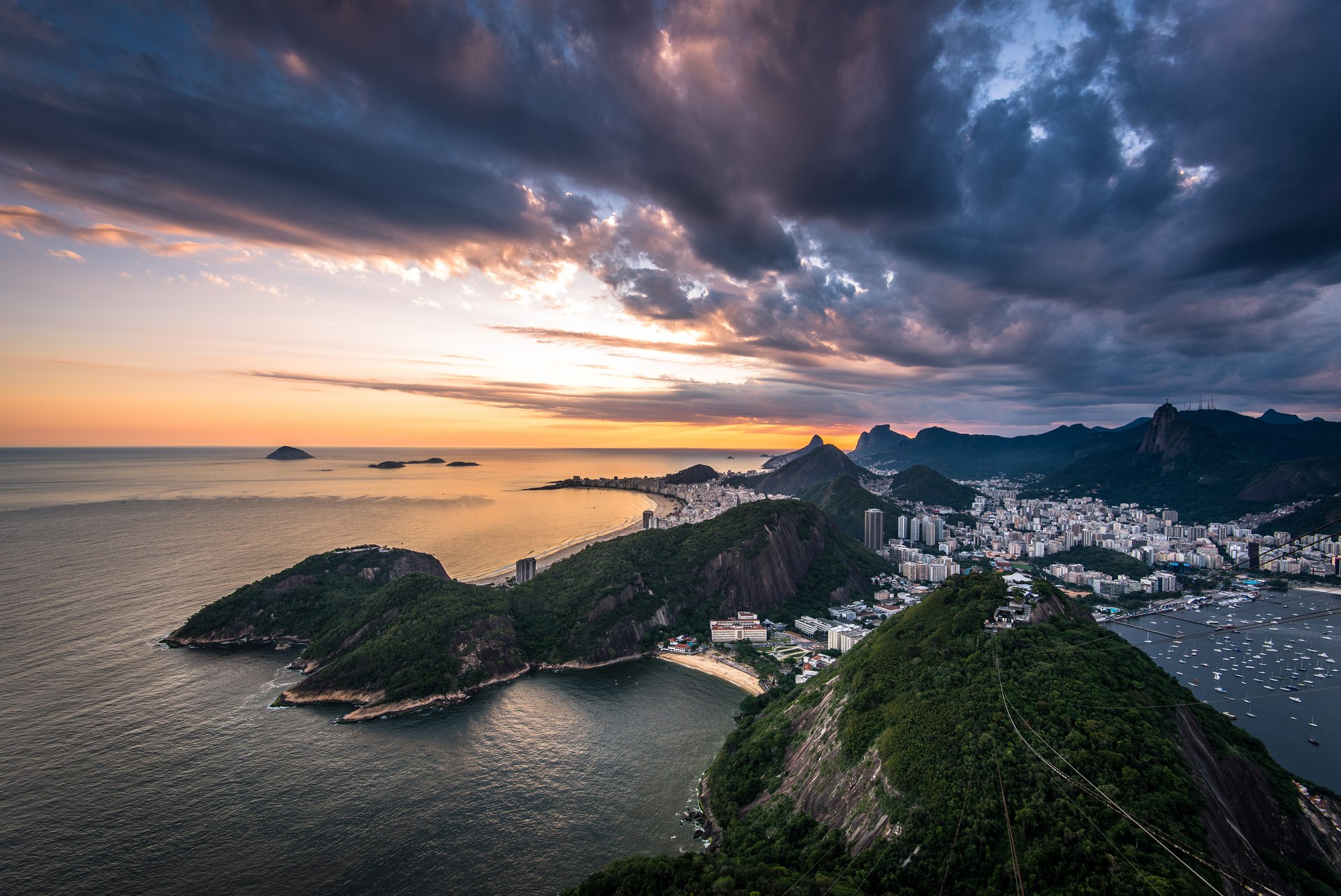 Vista panorâmica do Rio de Janeiro ao pôr do sol, com mar, praias, montanhas cobertas de vegetação e a cidade ao fundo.